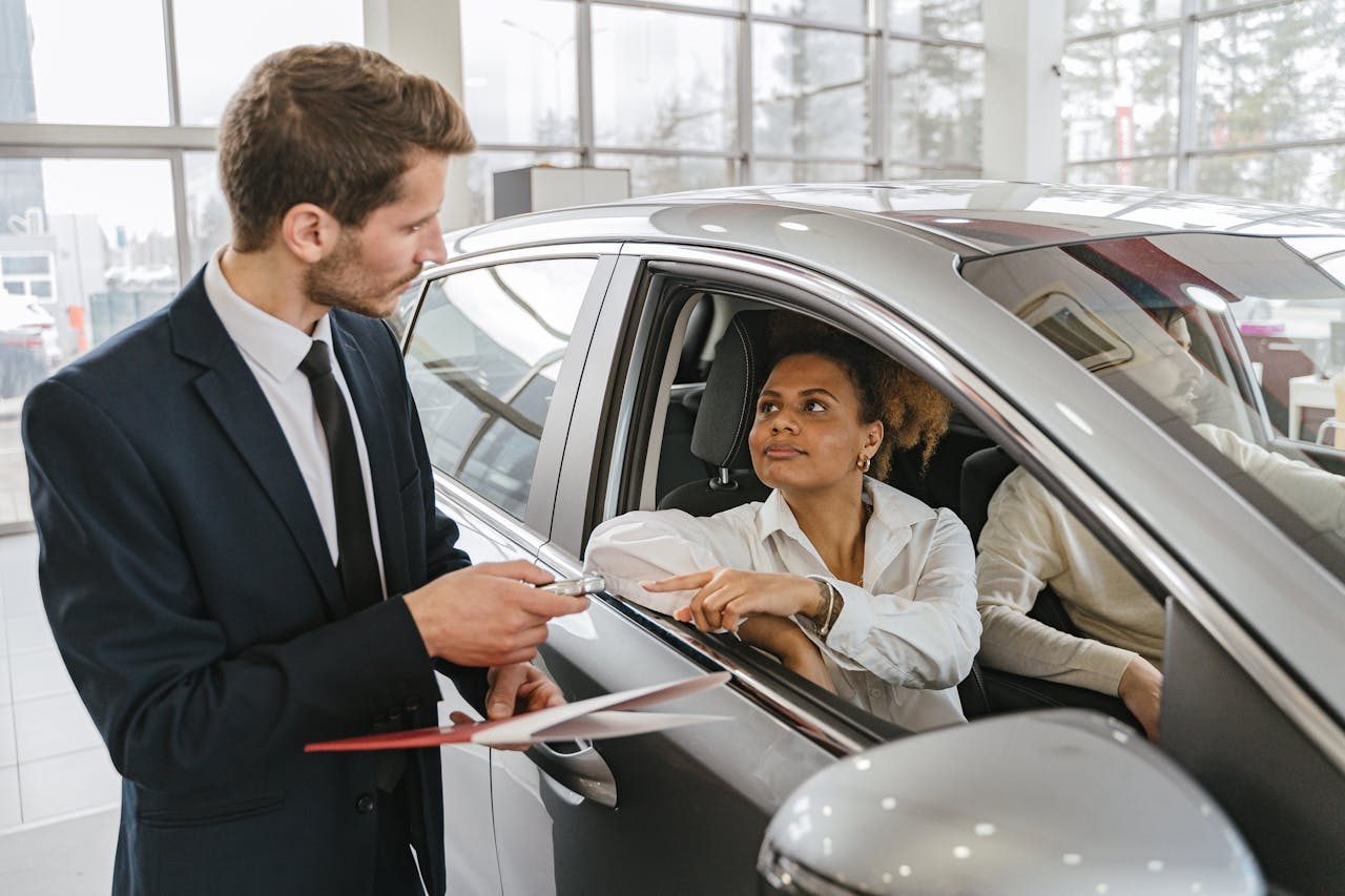team-03 A car dealer hands keys to a woman sitting inside a vehicle in a car showroom.